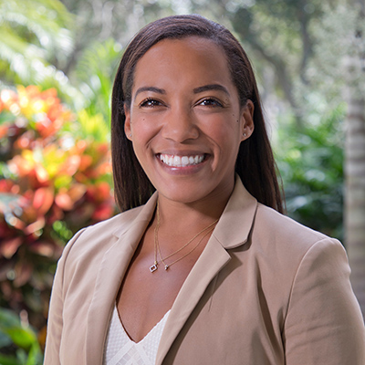 Portrait of a smiling woman in a tan suit and white blouse standing in front of green plants